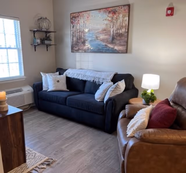 A cozy living room with a dark blue sofa adorned with white and striped pillows and a light throw blanket. Next to the sofa is a brown leather armchair with a white pillow and a red cushion. A small wooden side table with a lamp and a green plant is positioned between the sofa and armchair. On the wall above the sofa hangs a large framed painting of a serene river landscape with trees. A window with blinds lets in natural light, and a small shelf with decorative items is mounted on the adjacent wall. The floor is light wood, and a small wooden cabinet with a candle is partially visible.