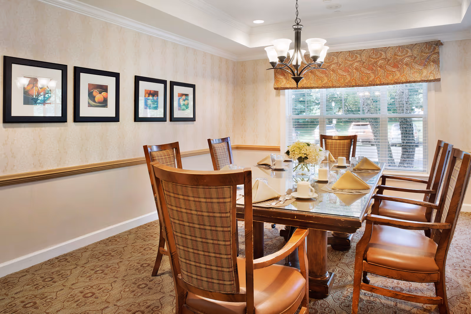 A dining room with a rectangular glass-top table set for six people. The table has folded napkins, cups, glasses, and a centerpiece with white flowers. The room features wooden chairs with cushioned seats and plaid backs, beige patterned wallpaper, a window with blinds and a decorative valance, and framed artwork on the wall. A chandelier with multiple lights hangs above the table.