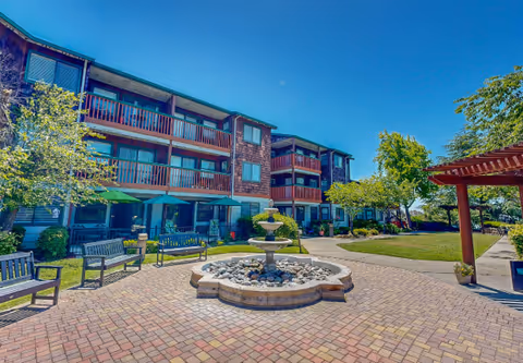 Courtyard of a senior living facility featuring a central fountain, benches, and a three-story building with balconies under a clear blue sky.