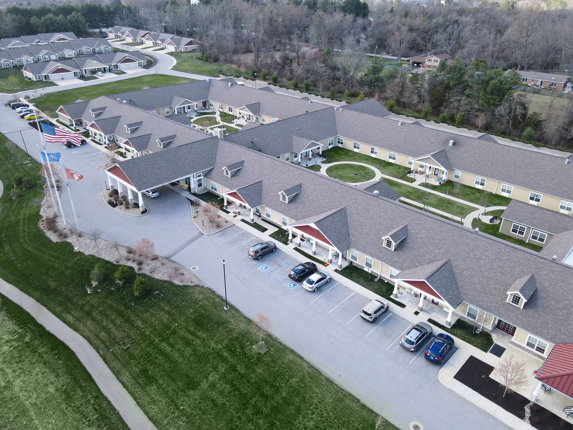 Aerial view of Gentry Park Senior Living facility showing multiple connected buildings with pitched roofs, parking spaces with several cars, landscaped green areas, walking paths, and flagpoles with flags near the entrance.