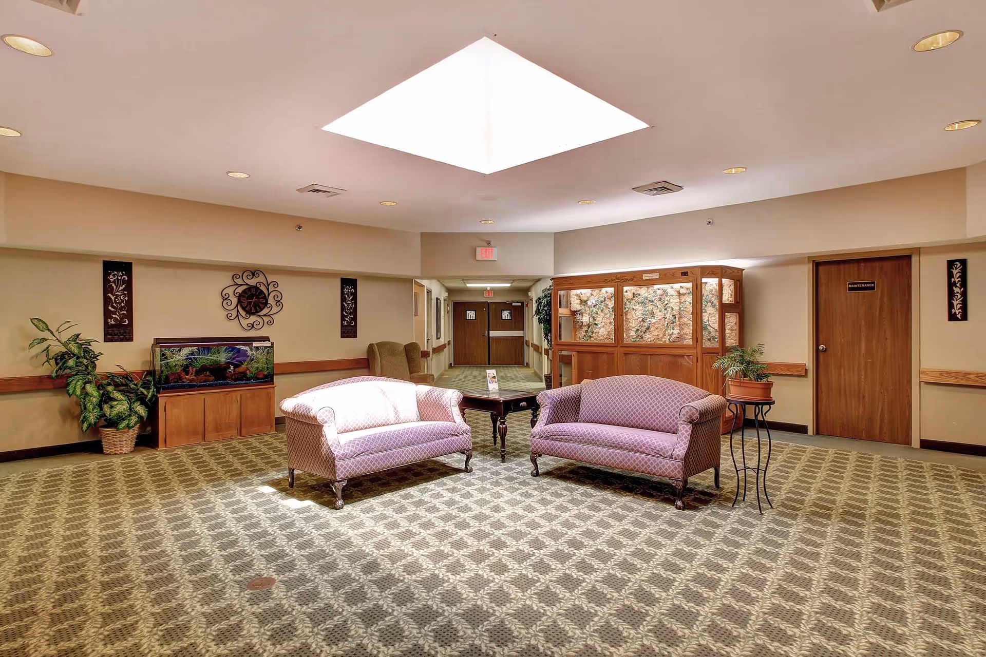 A well-lit common area in an assisted living facility featuring two patterned sofas facing each other with a small wooden table between them. The room has a patterned carpet, a skylight in the ceiling, a fish tank on a wooden stand, potted plants, and a wooden door labeled 'Maintenance'. The walls are decorated with framed art and a clock.