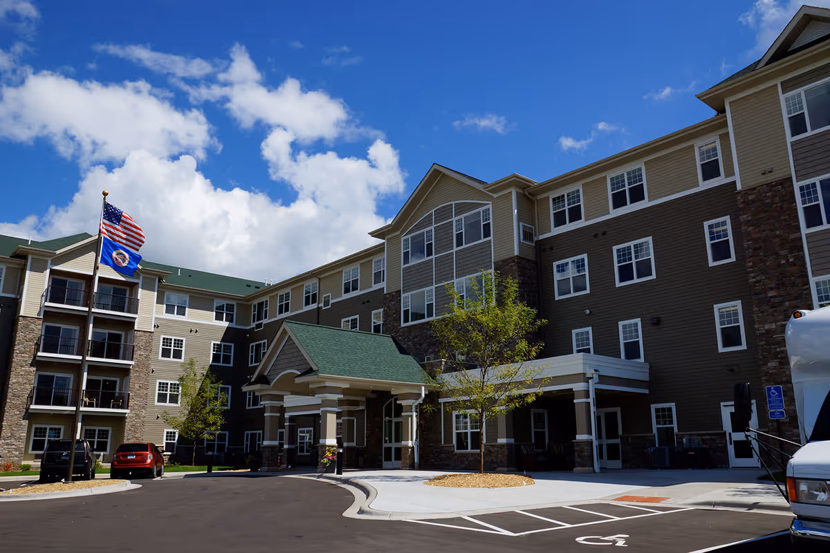 Exterior view of a multi-story senior living facility building with a covered entrance, several windows, and a green roof. There are two flags on a flagpole, a few parked cars, a tree, and a clear blue sky with some clouds.