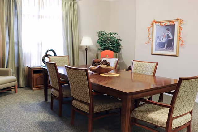 A dining room with a wooden table surrounded by six upholstered chairs. The room has light-colored walls, a window with green curtains, a floor lamp, a potted plant, and a framed picture on the wall. There is a decorative bowl with pine cones on the table.