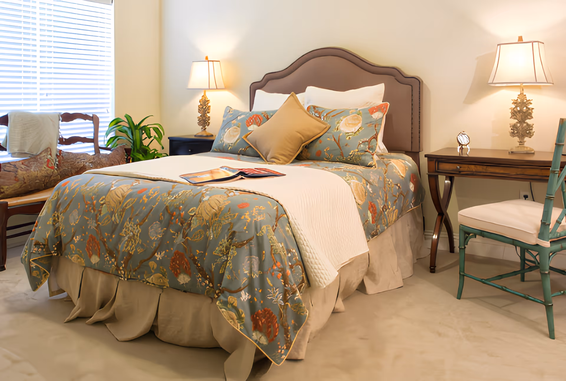 A cozy bedroom with a floral-covered bed, nightstands with lamps, chairs, and a window with blinds.