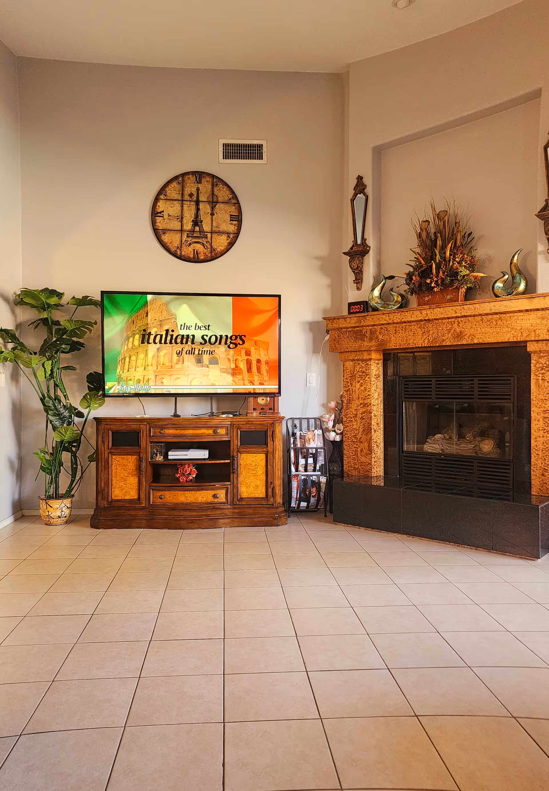 A cozy living room area featuring a wooden TV stand with a flat-screen TV displaying 'the best italian songs of all time' with an image of the Colosseum and the Italian flag. To the right is a decorative fireplace with a floral arrangement and ornamental pieces on the mantel. A round wall clock with the Eiffel Tower design hangs above the TV. A tall green plant is placed to the left of the TV stand. The floor is tiled with light-colored tiles.