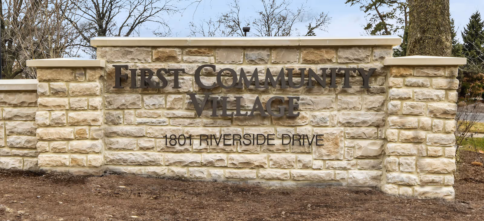 Stone sign at the entrance of a facility with the text 'First Community Village' and the address '1801 Riverside Drive'. The sign is made of light-colored stone blocks with dark metal letters. Trees and a road are visible in the background.
