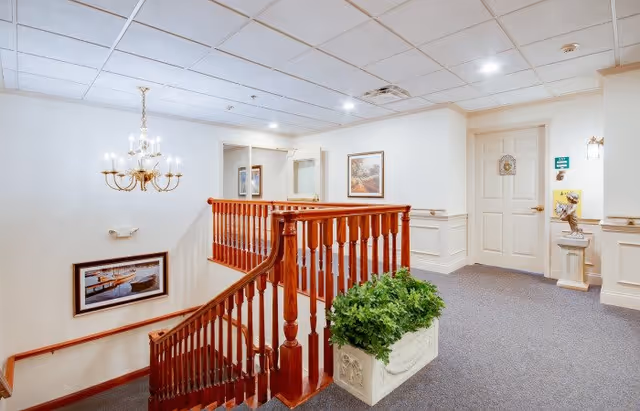 Interior view of a senior living facility hallway with a wooden staircase railing, a chandelier hanging from the ceiling, framed artwork on the walls, a green potted plant in a white planter, and a closed door at the end of the hallway.