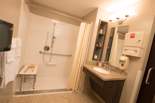 Accessible bathroom with a walk-in shower and bench, grab bars, sink, mirror and towel dispenser.