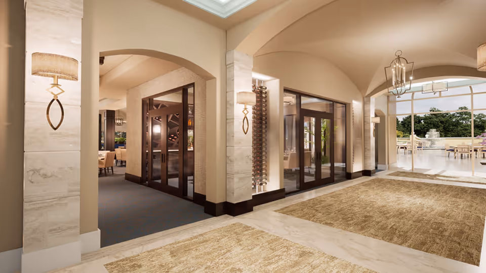 Spacious, well-lit interior hallway with arched ceilings and beige walls. The floor is covered with large marble tiles and two large beige rugs. Wall sconces with lamps provide warm lighting. Glass doors lead to a dining area on the left and an outdoor patio with tables and a fountain visible through large windows on the right.