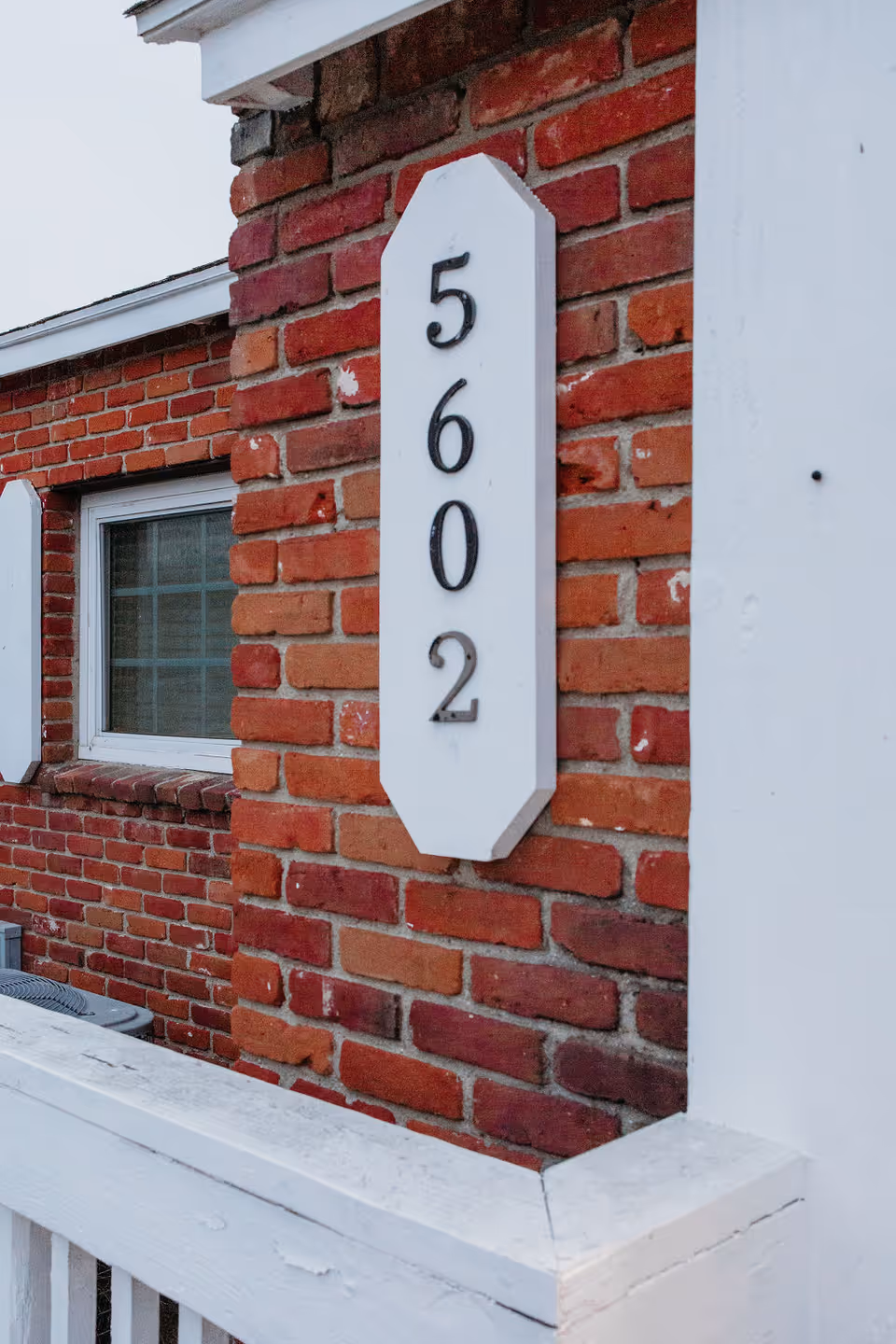 Brick building exterior showing a white vertical house number plaque reading 5602 beside a window and a white railing.