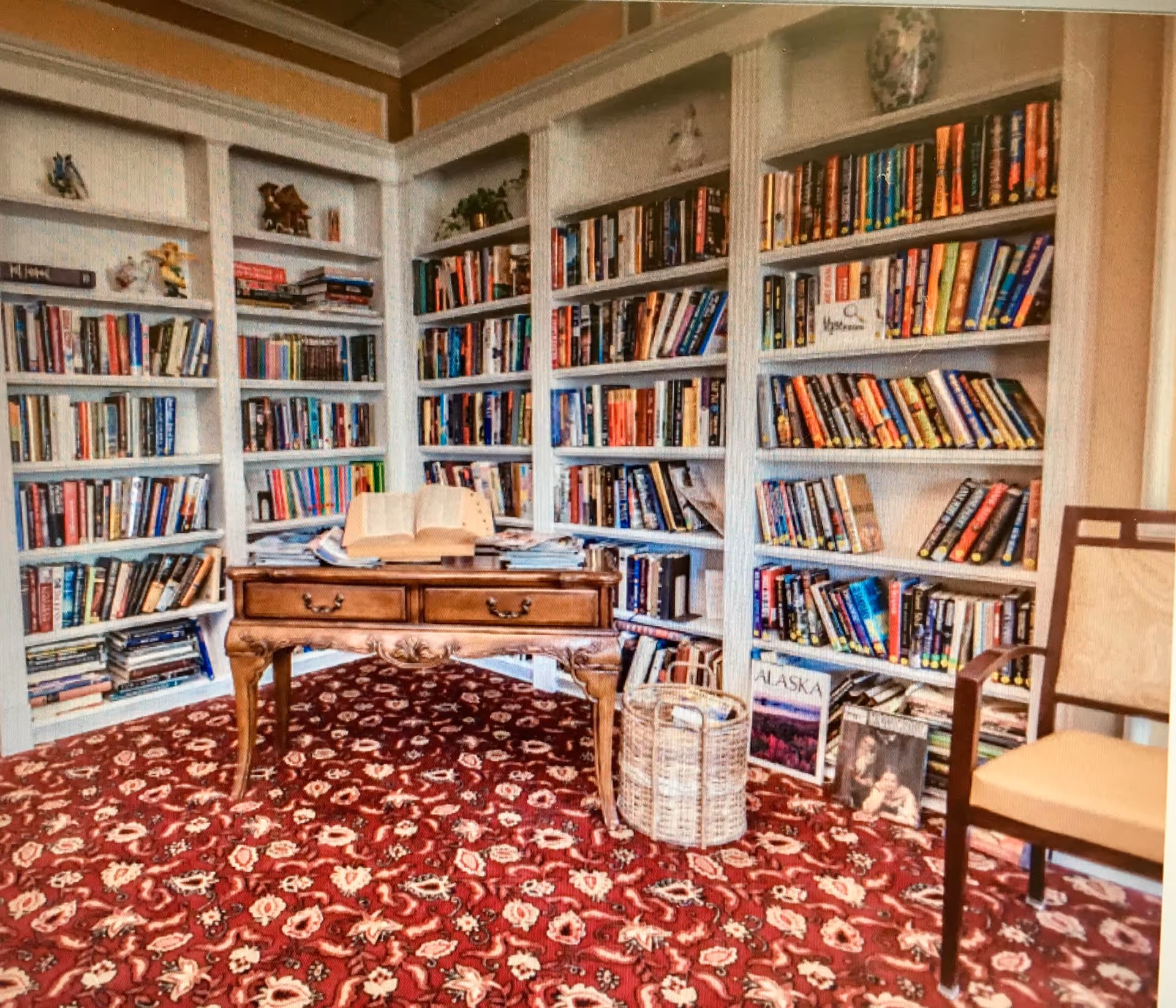 Cozy library room with white built-in bookshelves filled with books, a wooden table with an open book on top, a wicker basket, and a chair with a beige cushion. The floor is covered with a red patterned carpet.