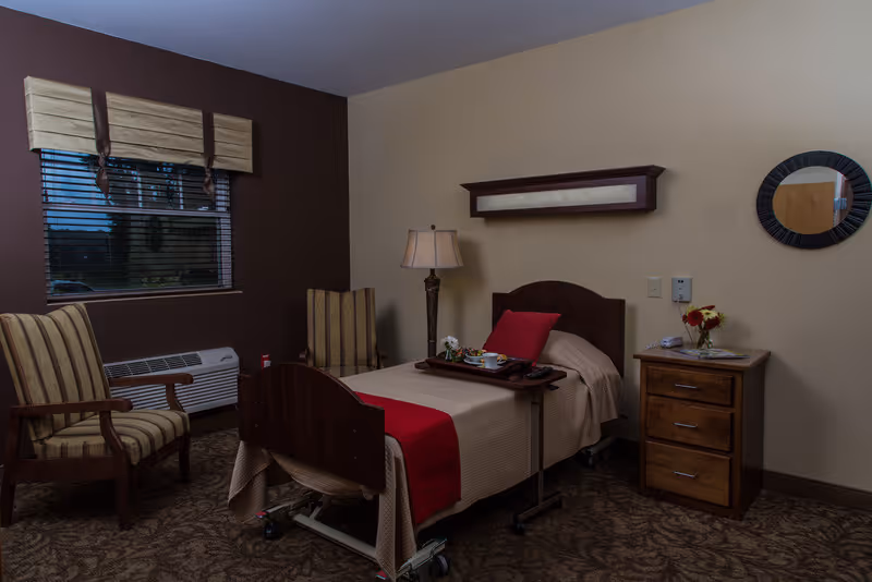 A neatly arranged bedroom in a senior living facility featuring a single hospital-style bed with beige bedding and a red throw blanket. There is a tray with a tea set on the bed. Two striped armchairs are positioned near a window with blinds and a beige valance. A wooden nightstand with drawers holds a vase of flowers and some items. A floor lamp stands beside the bed, and a round mirror hangs on the wall.