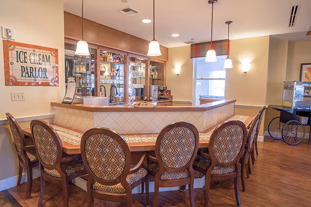 Interior view of an ice cream parlor area with a U-shaped counter surrounded by eight patterned chairs. The counter has a tiled front and wooden top, with pendant lights hanging above. Behind the counter is a glass cabinet displaying various items, and a sign on the wall reads 'Ice Cream Parlor'. The room has warm lighting and wooden flooring.
