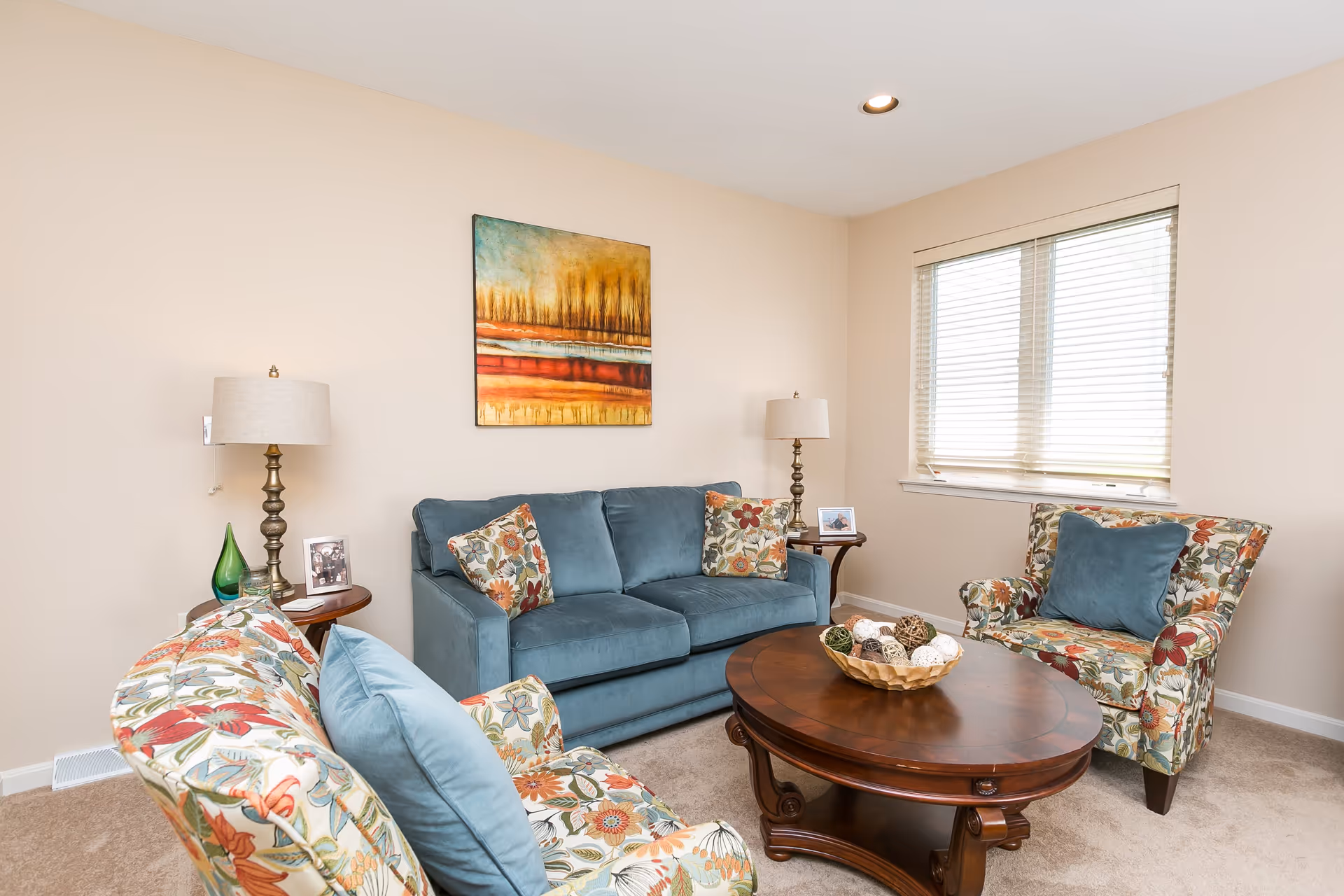 A cozy living room with a blue sofa and two floral-patterned armchairs surrounding a wooden coffee table. The coffee table has a decorative bowl with assorted decorative balls. There are two side tables with lamps and framed photos, and a large window with blinds letting in natural light. A colorful abstract painting hangs on the beige wall behind the sofa.