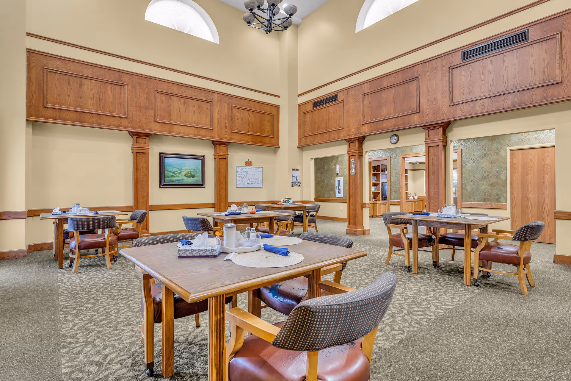 Dining room with multiple wooden tables and cushioned chairs set for meals beneath high ceilings and wood-paneled columns.