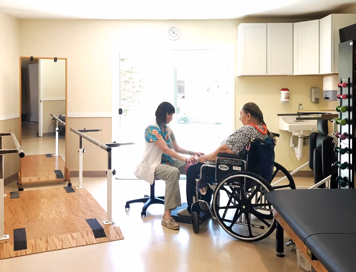 A healthcare worker in a white vest and floral shirt kneels in front of an elderly woman in a wheelchair, holding her hands in a bright therapy room with parallel bars, a large mirror, a treatment table, and cabinets with medical supplies.
