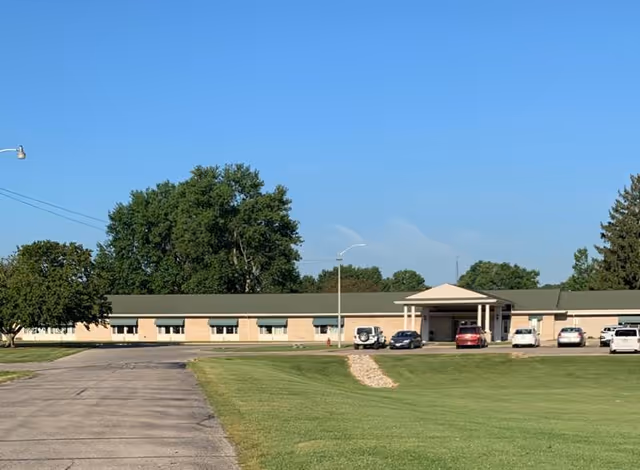 Exterior view of a single-story building with a green roof and beige walls, surrounded by a large grassy area and several parked cars in front under a clear blue sky.