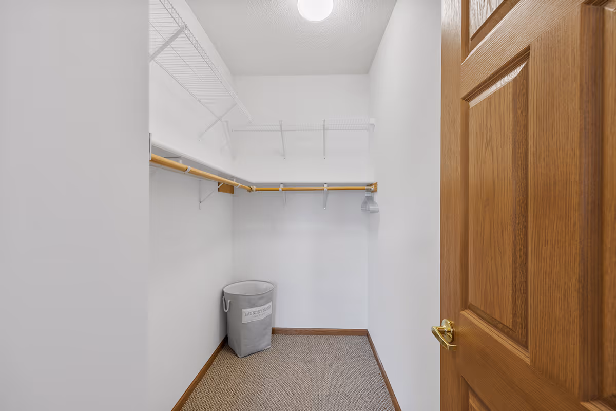 Empty walk-in closet with white walls, beige carpet, wooden door with brass handle, wire shelving, wooden hanging rods, and a gray laundry basket labeled 'Laundry Basket'.
