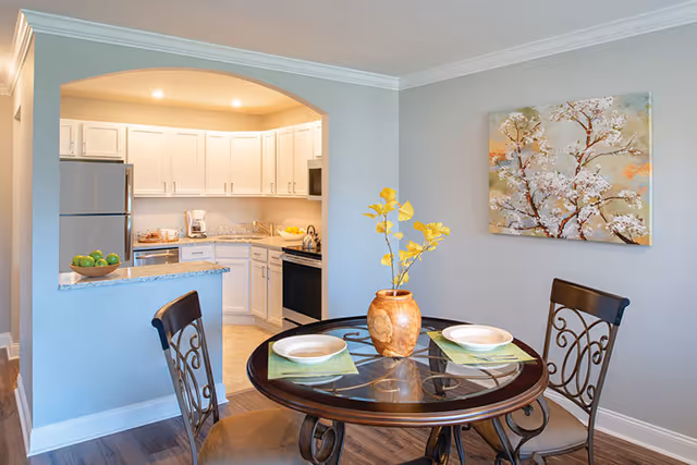 Small dining area with a round glass-top table set for two and a vase of yellow flowers, adjacent to an open kitchen with white cabinets.