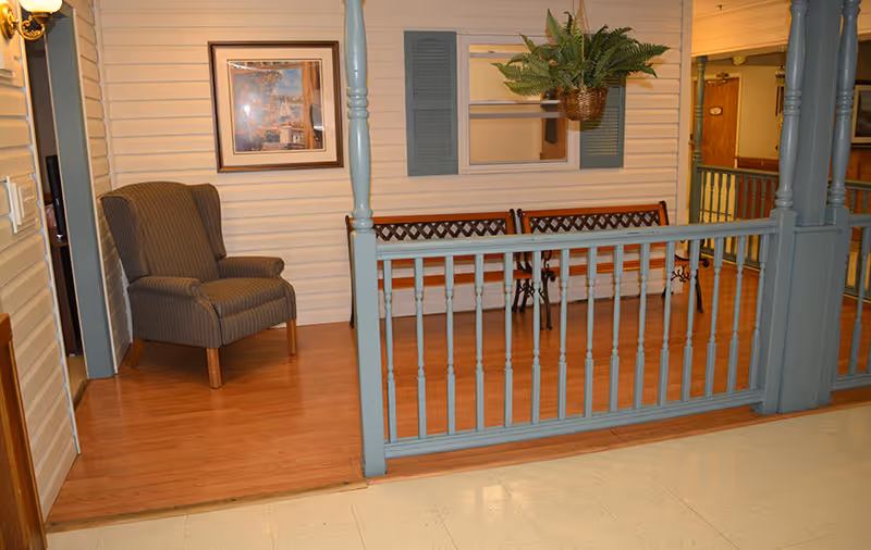 Indoor common area styled like a porch with a blue railing, a bench, an armchair, framed art and a hanging plant.