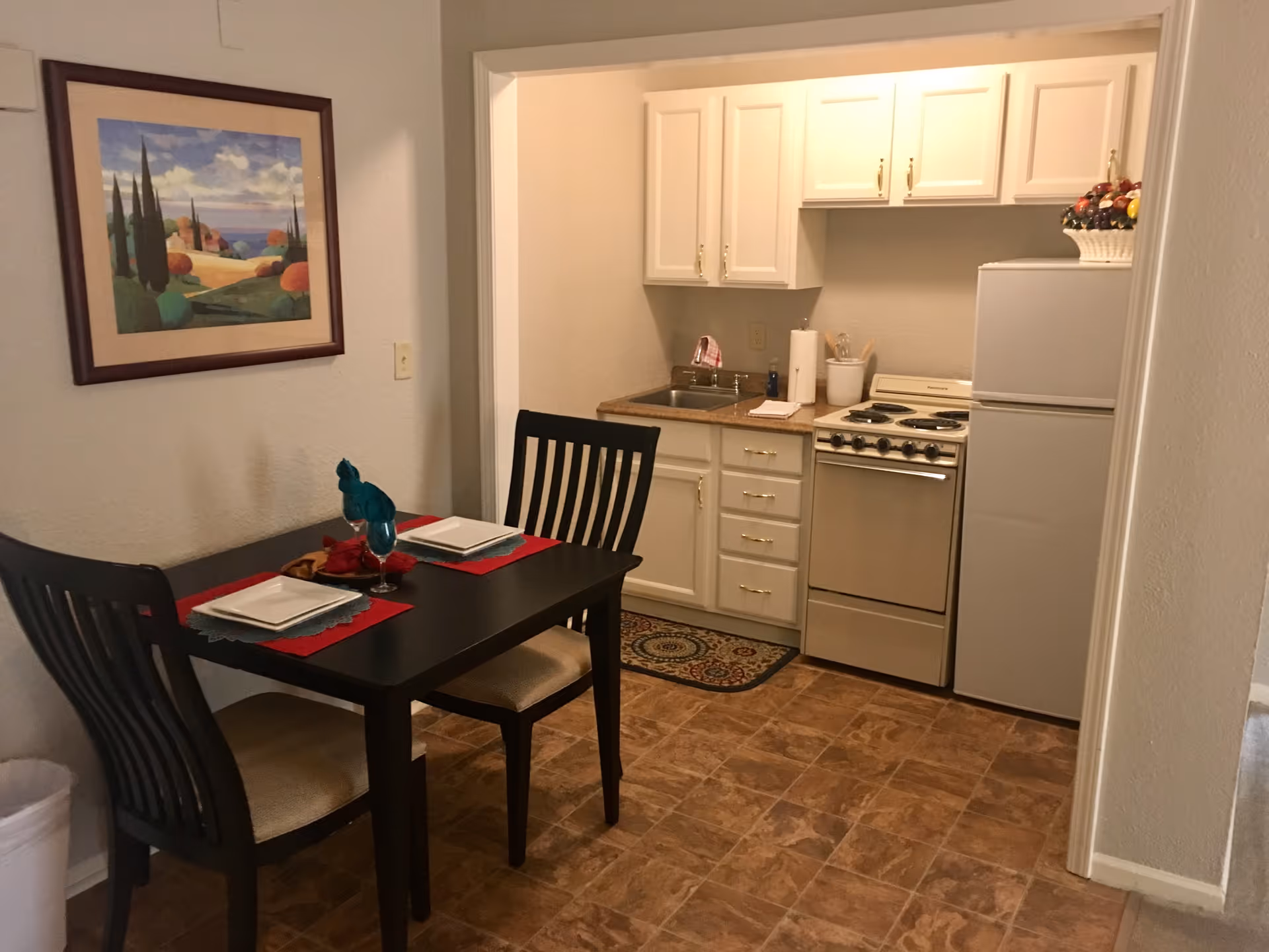 Small apartment kitchenette with a dining table set for two, stove, refrigerator, sink and wall art.