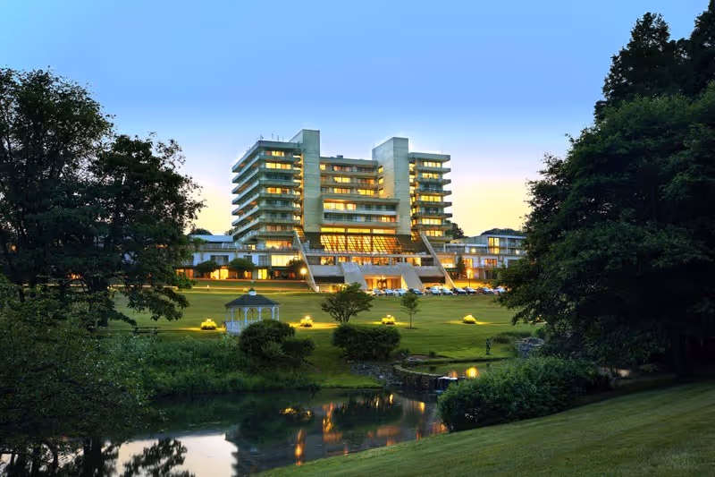 A large multi-story building with many windows lit up at dusk, surrounded by well-maintained green lawns, trees, a small pond, and a white gazebo in the foreground.