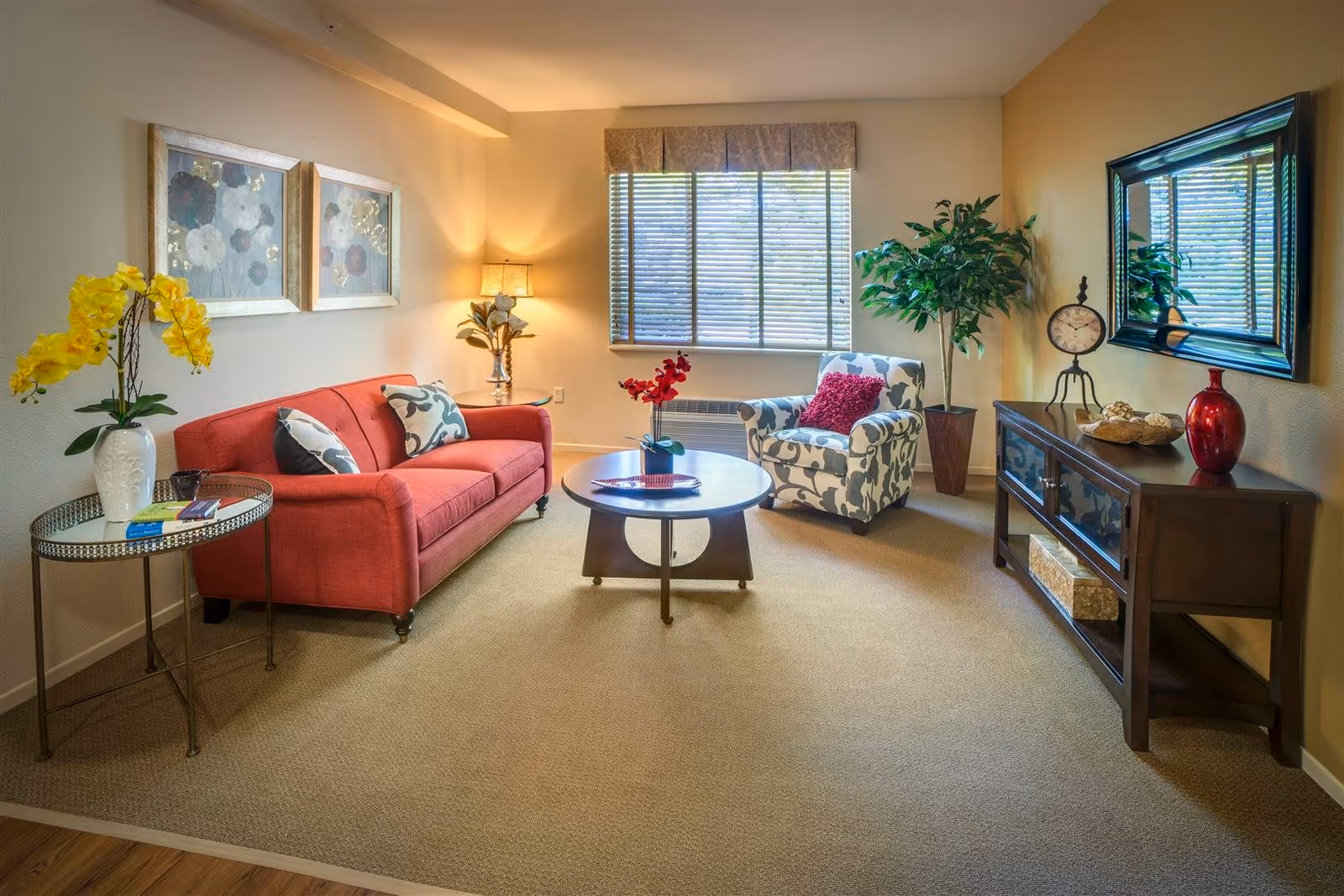 A cozy living room with a red sofa adorned with patterned pillows, a round wooden coffee table with a small plant, and a patterned armchair with a red cushion. The room features a window with blinds, a side table with a lamp and decorative plant, a console table with a clock, red vase, and decorative items, and two framed floral paintings on the wall.