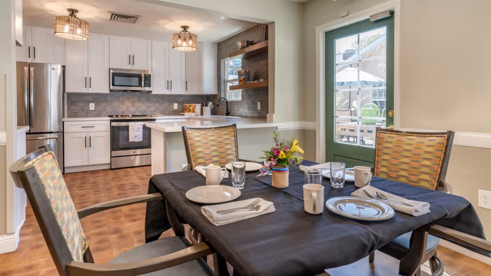A dining area with a table set for four people, featuring plates, cups, glasses, and napkins on a black tablecloth. The chairs have patterned upholstery. In the background, there is a modern kitchen with white cabinets, stainless steel appliances including a refrigerator, oven, and microwave, and two ceiling light fixtures. A green door with glass panels leads outside to a patio area with an umbrella and outdoor furniture.