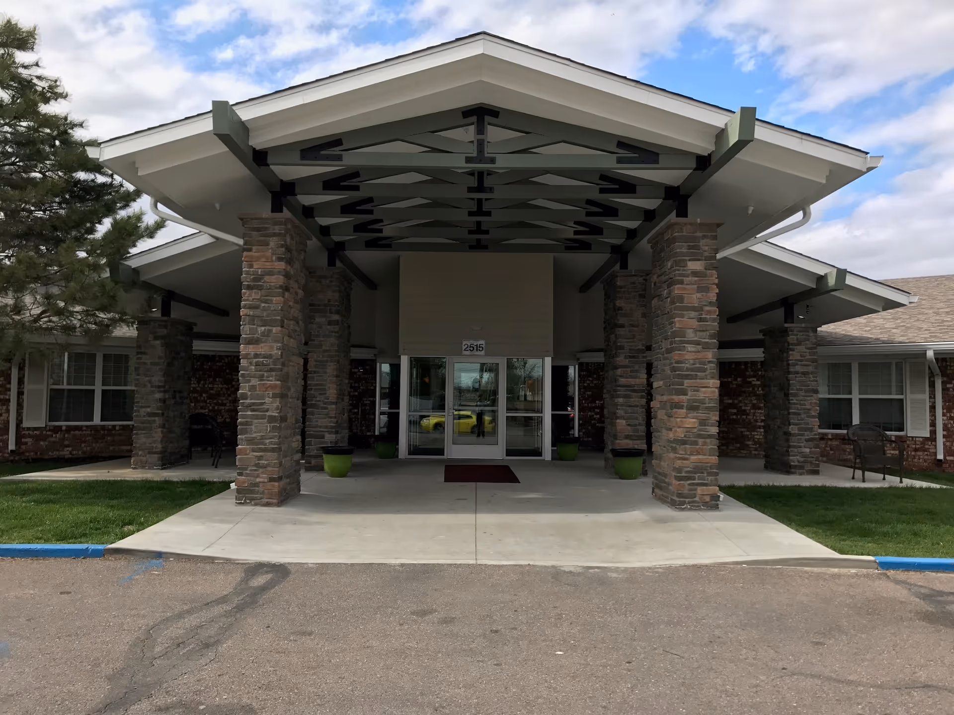Front entrance of a building with a covered driveway supported by stone pillars. The building has large glass double doors and windows on either side. There are green plants in pots near the entrance and a small patch of grass on both sides of the driveway. The sky is partly cloudy.