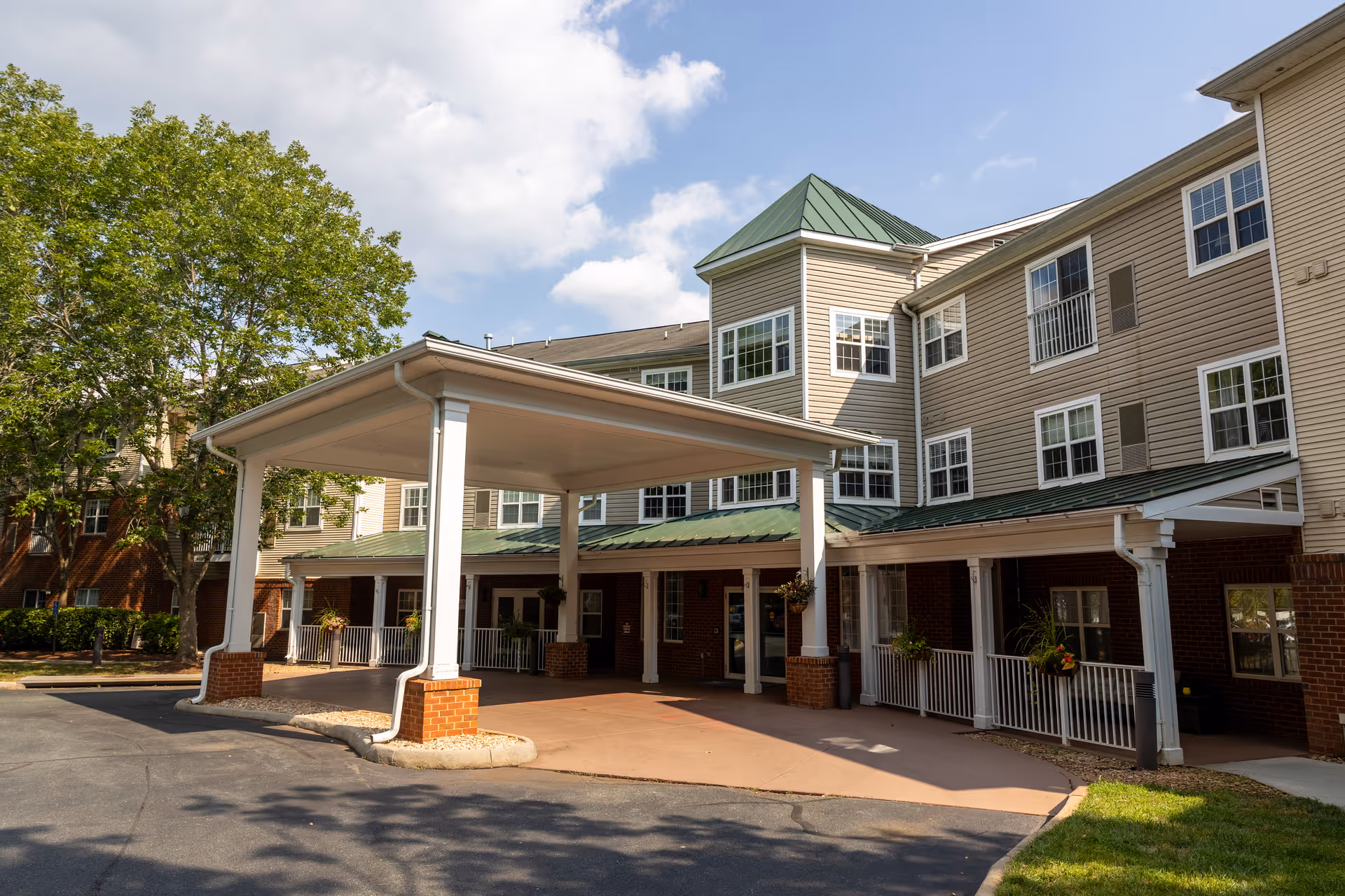 Exterior view of Commonwealth Senior Living At Charlottesville showing the front entrance with a covered drop-off area, beige siding, green metal roofs, multiple windows, and a large tree to the left under a partly cloudy sky.