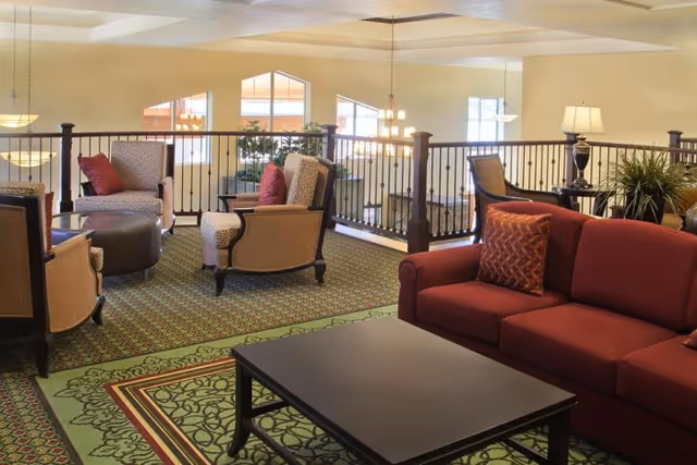 A cozy living room area with a red sofa, patterned armchairs, a black coffee table, and decorative pillows. The room has a green patterned carpet and is well-lit with hanging and table lamps. There is a railing overlooking a lower level with large windows in the background.