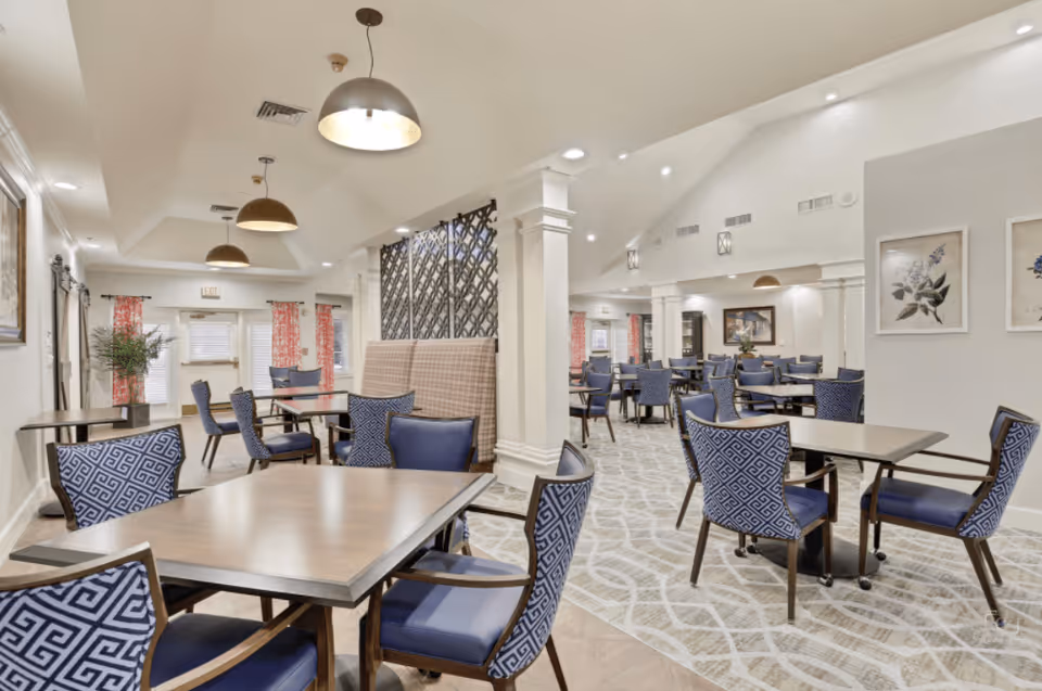 A spacious dining room with multiple tables and blue patterned chairs. The room features high ceilings with recessed lighting and hanging pendant lights. There are decorative columns, framed botanical artwork on the walls, and large windows with red patterned curtains allowing natural light to enter.