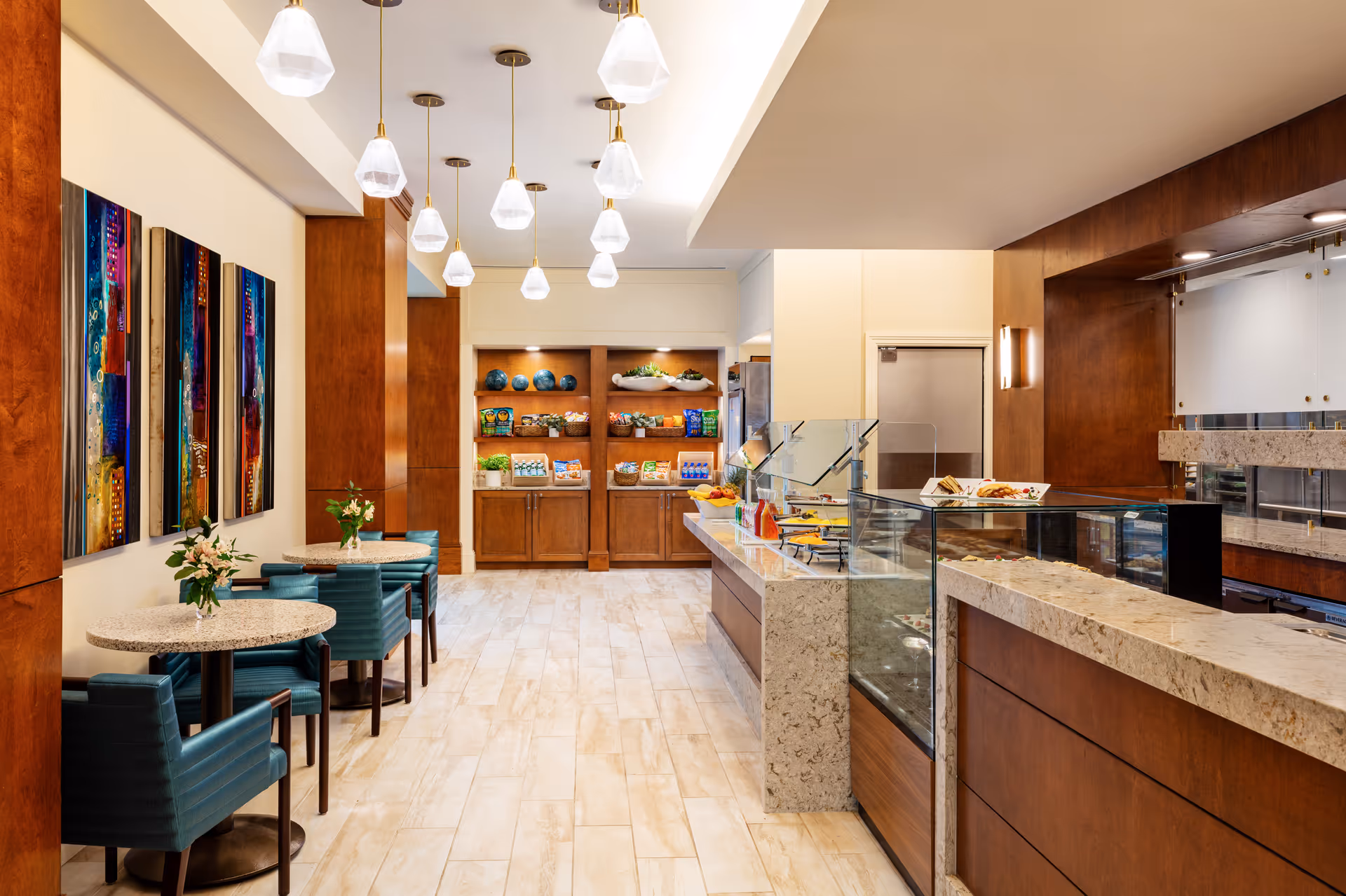 A modern dining area with small round tables and teal upholstered chairs. The room features a buffet counter with a glass sneeze guard displaying food items, and wooden shelves stocked with snacks and decorative items. Pendant lights hang from the ceiling, and colorful abstract artwork is displayed on the wall.