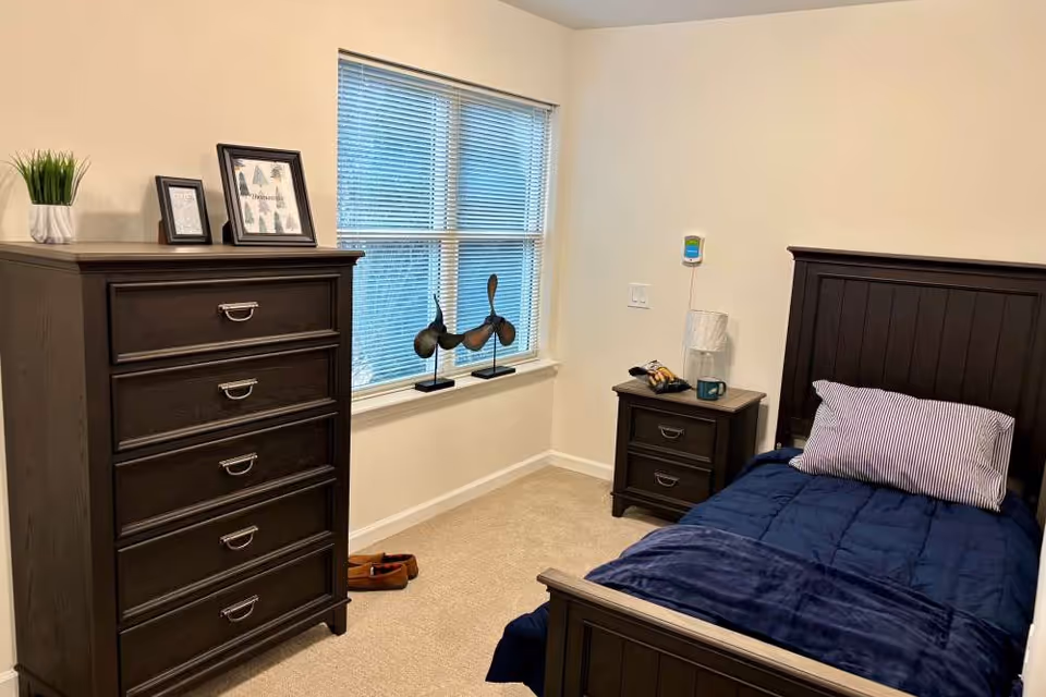 A small bedroom with a dark wooden bed frame and matching nightstand and dresser. The bed is made with a navy blue comforter and a striped pillow. There is a window with blinds letting in natural light, and decorative items including framed pictures and sculptures are placed on the dresser and windowsill.