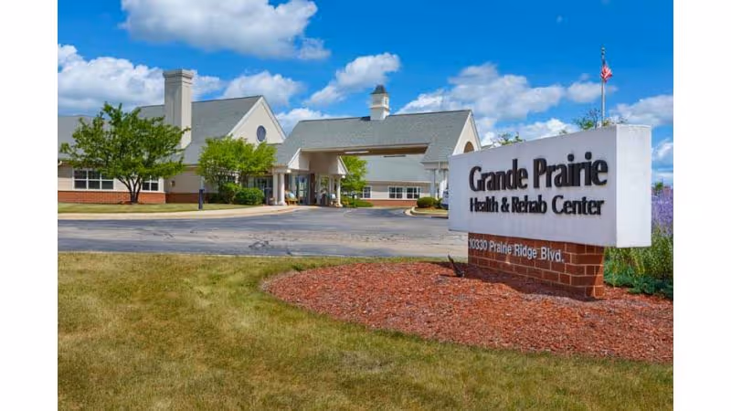 Exterior front entrance of Grande Prairie Health & Rehab Center with driveway, landscaping, and a large roadside sign.