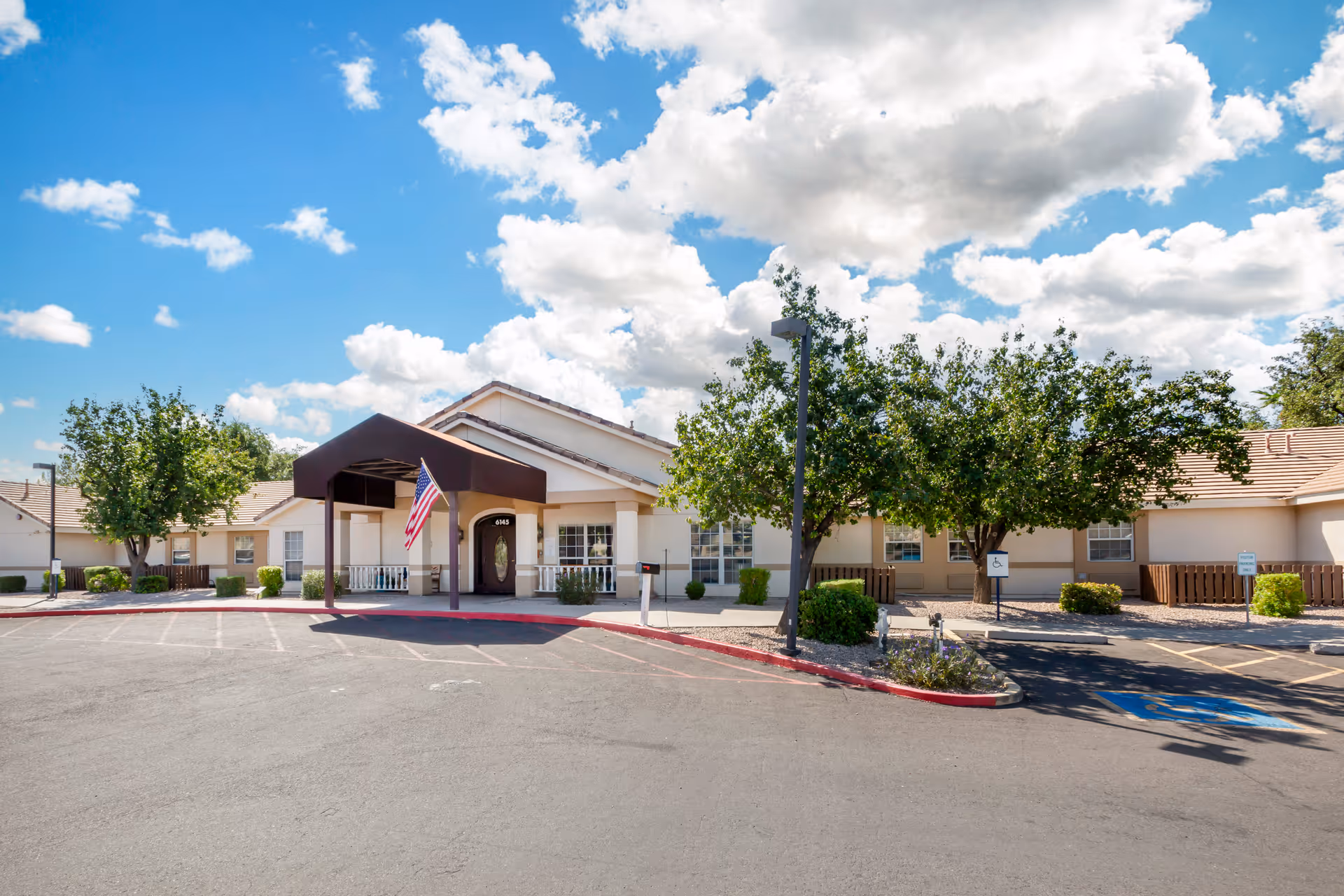 Exterior view of a single-story building with a covered entrance and an American flag hanging from the canopy. The building is surrounded by trees and bushes, with a parking lot in front including a handicapped parking space. The sky is partly cloudy with blue patches.