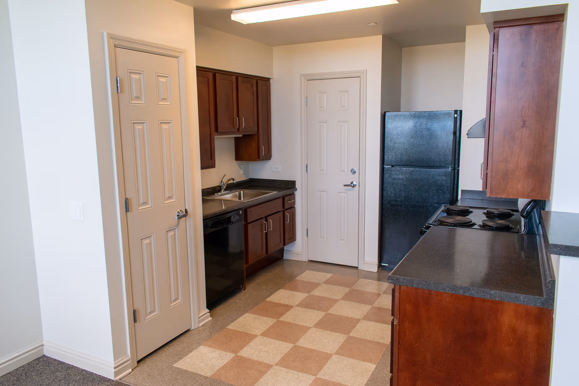 A kitchen with wooden cabinets, a black refrigerator, a black stove with four burners, a double sink, and a dishwasher. The floor has a checkered pattern with beige and light brown tiles. There are two closed doors, one on the left and one straight ahead.