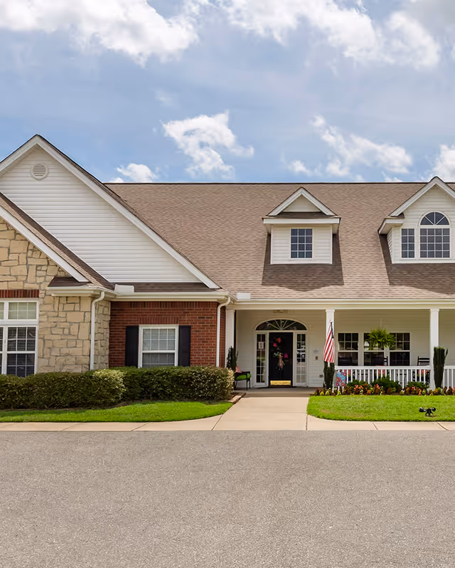 Front exterior of a residential-style building with a covered entrance, porch, American flag, and manicured lawn and shrubs.