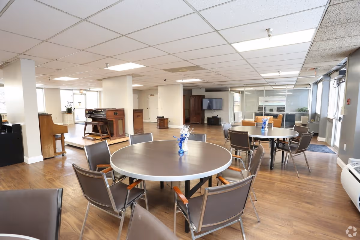 A spacious common area with round tables and chairs arranged for seating. The room features wooden flooring, a piano and organ on a small raised platform, a flat-screen TV mounted on the wall, and large windows letting in natural light. There are some small decorative flower arrangements on the tables.