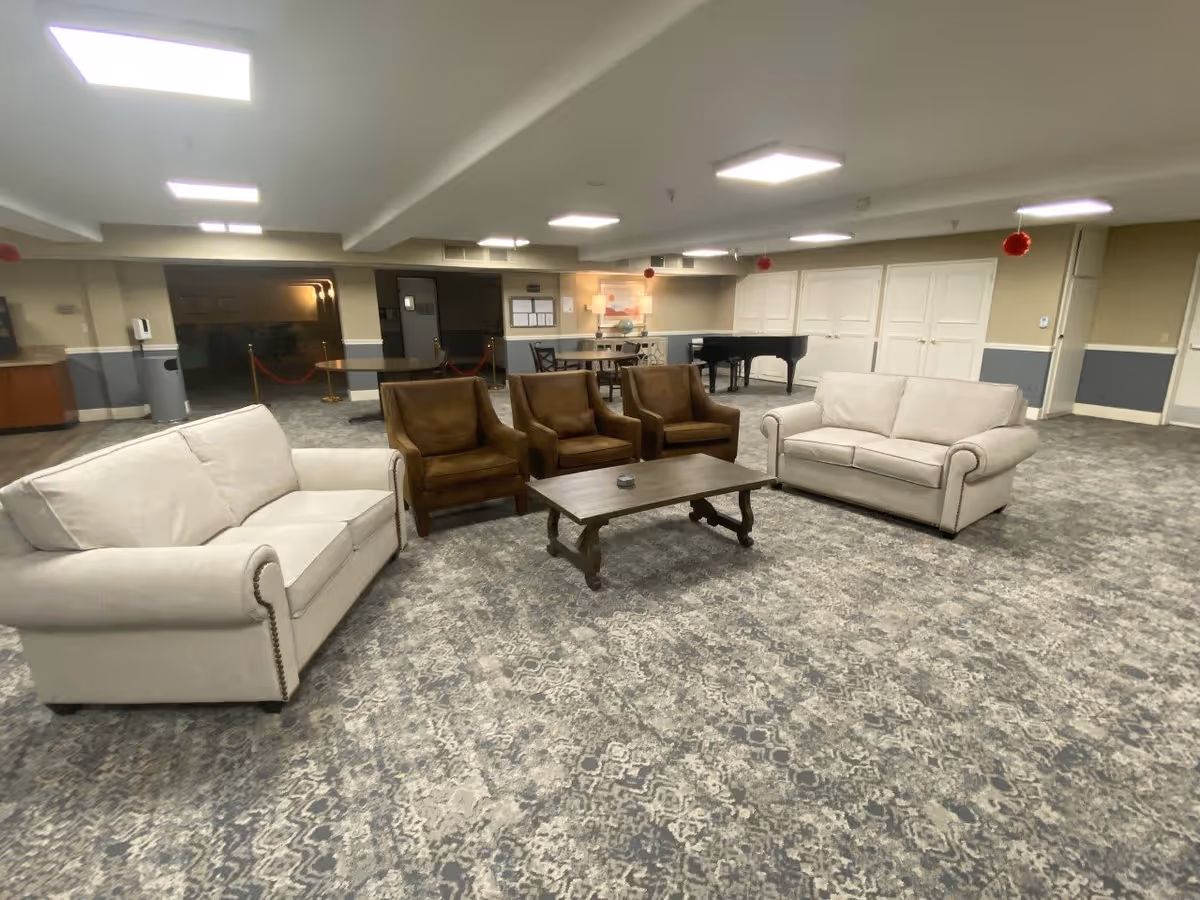 A spacious senior living facility common area with two beige sofas and three brown armchairs arranged around a wooden coffee table on a patterned carpet. In the background, there is a black grand piano, a table with chairs, and several closed white doors. The room is well-lit with ceiling lights and decorated with small red hanging ornaments.