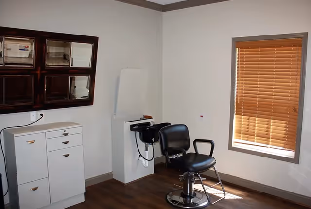 Small salon room with a black styling chair, shampoo bowl, cabinets, and a window with wooden blinds.