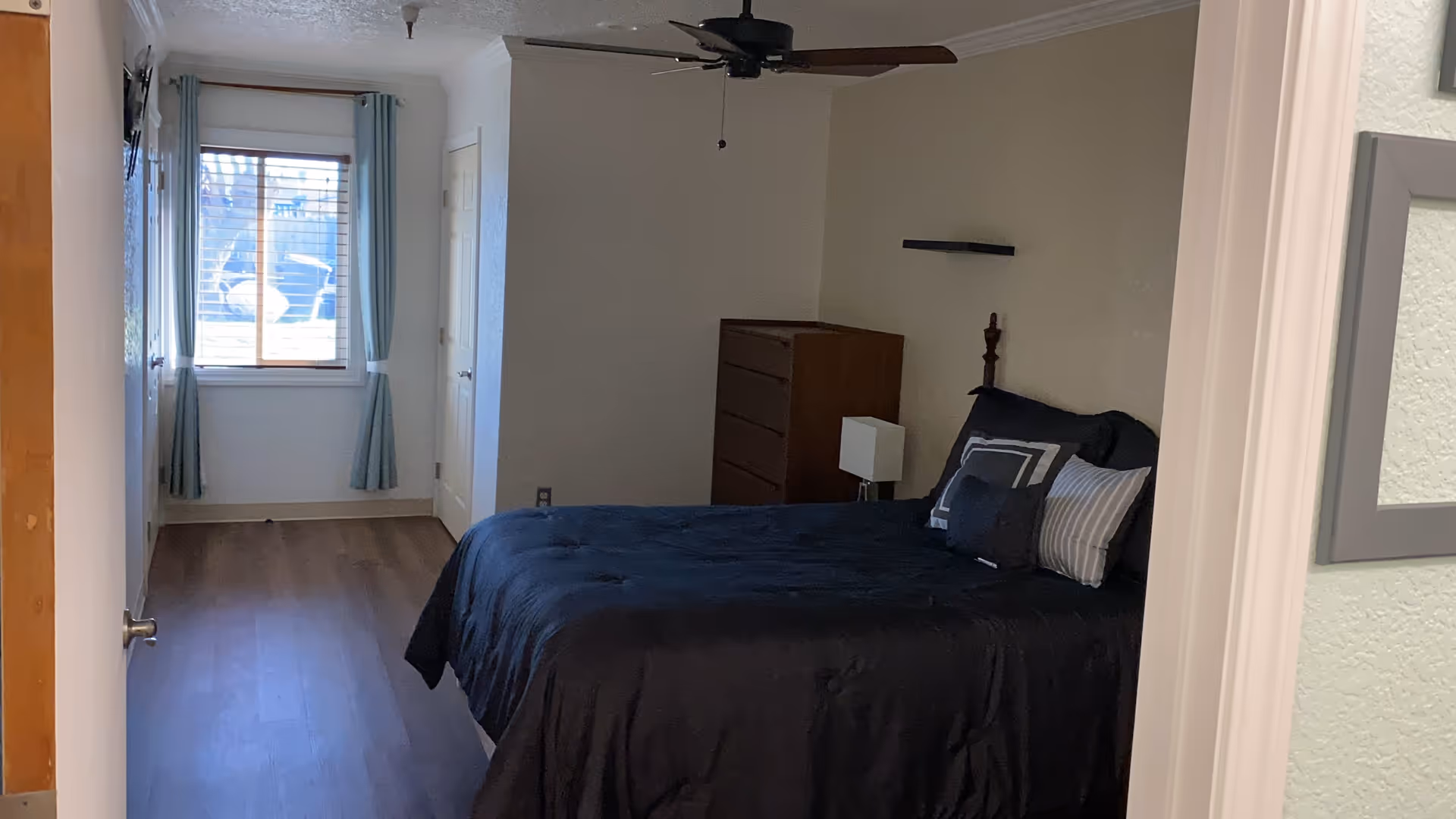 A bedroom with a dark bedspread on a bed, several pillows, a wooden dresser, a small lamp on a nightstand, a ceiling fan, and a window with light blue curtains letting in natural light.