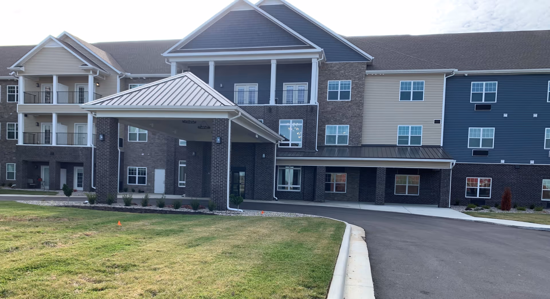 Front exterior of a three-story senior living building with a covered porte-cochere, driveway, lawn, and multiple balconies.
