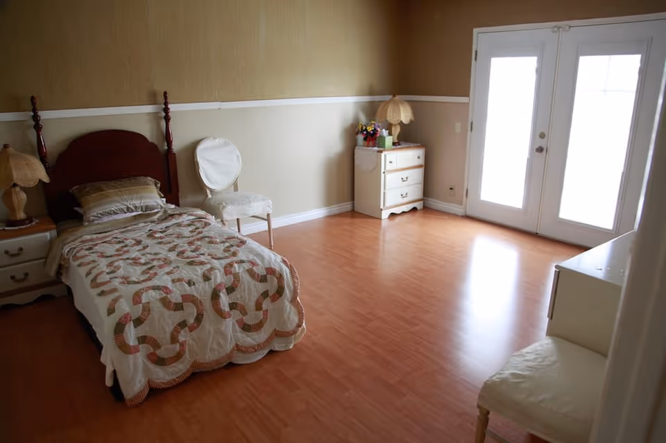 A simple bedroom with a single bed covered with a patterned quilt, two white chairs, two nightstands with lamps, a small dresser with flowers and a lamp, and double glass doors letting in natural light. The room has wooden flooring and beige walls with a white trim.