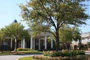 Front entrance of a brick retirement community building with white columns, landscaped flower beds, and trees.