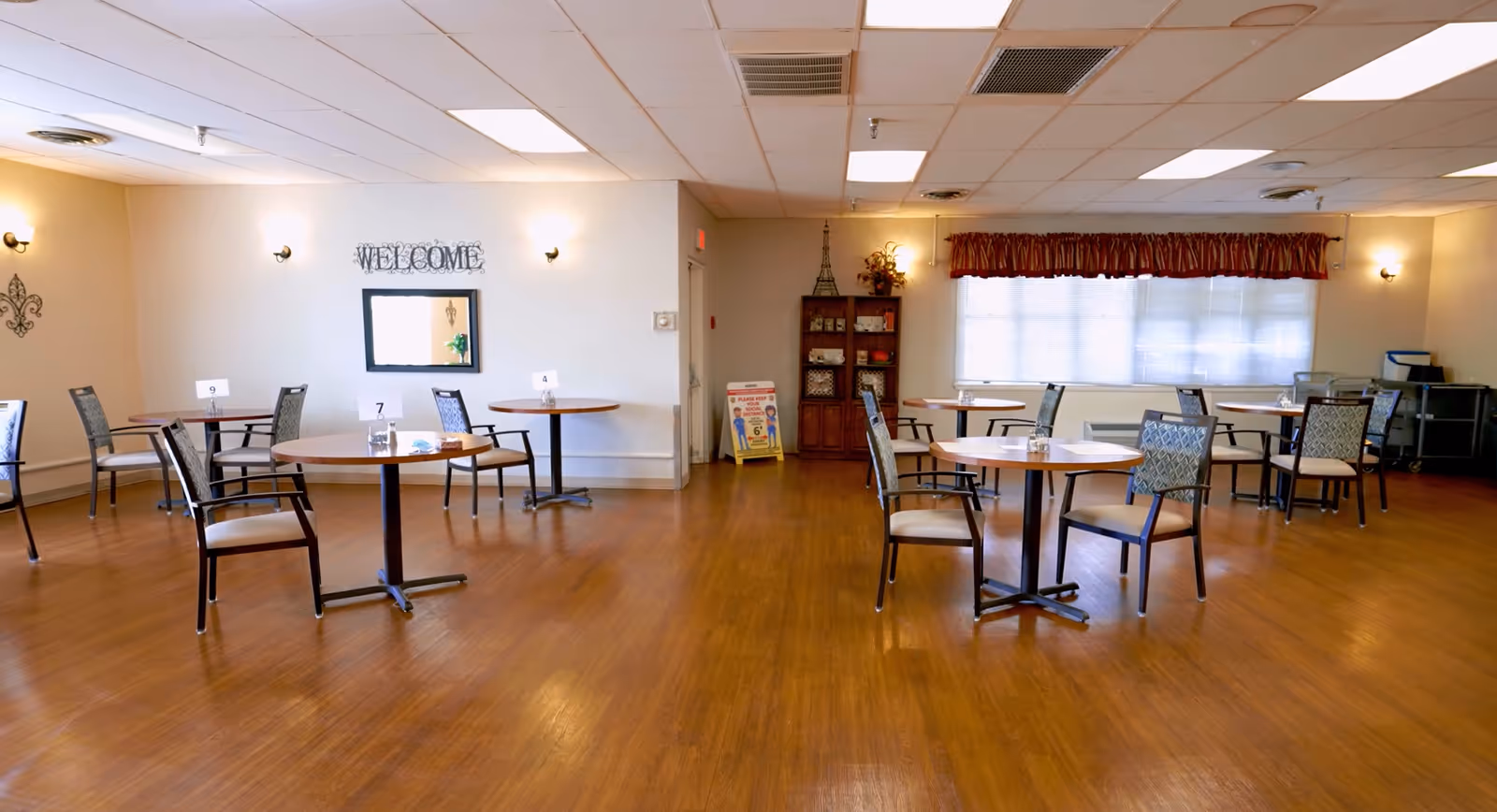 A spacious dining room with several round tables and chairs arranged neatly on a wooden floor. The walls are light-colored with wall sconces providing soft lighting. A mirror with the word 'WELCOME' above it is mounted on one wall. There is a window with a red valance curtain, a wooden cabinet with decorative items, and a small sign near the back wall.