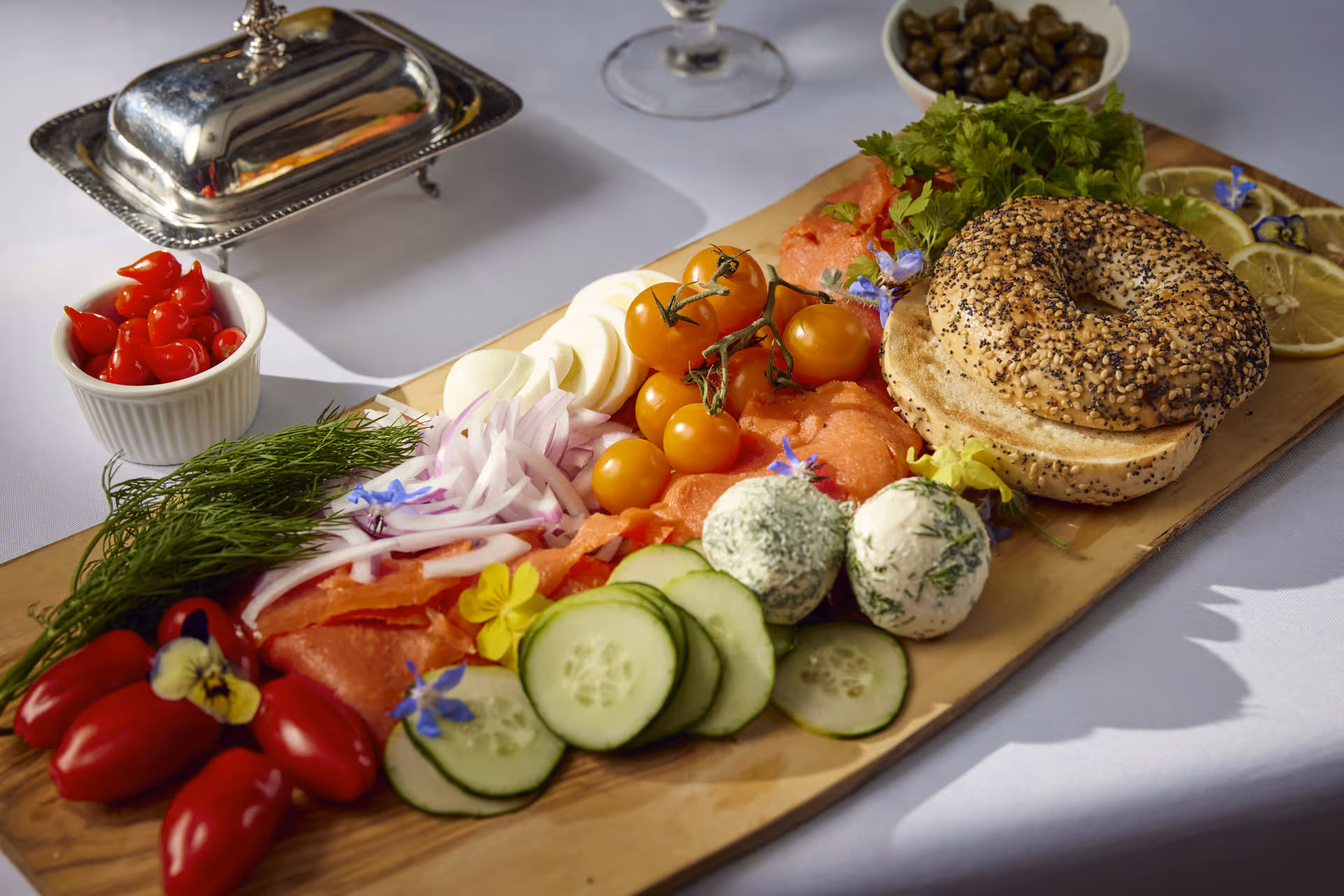 A wooden serving board with an assortment of fresh food including an everything bagel, cherry tomatoes on the vine, sliced cucumbers, red onions, smoked salmon, two herb-coated cheese balls, fresh dill, and small red peppers. There are also lemon slices and a small bowl of capers on the side, all placed on a white tablecloth.