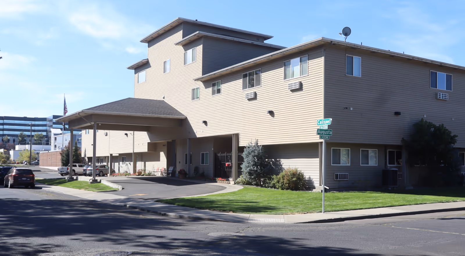 Exterior view of a multi-story beige building with a covered entrance and several windows, situated on a street corner with green grass and a few bushes around. There are cars parked along the side and a street sign showing Calispel and Augusta streets.