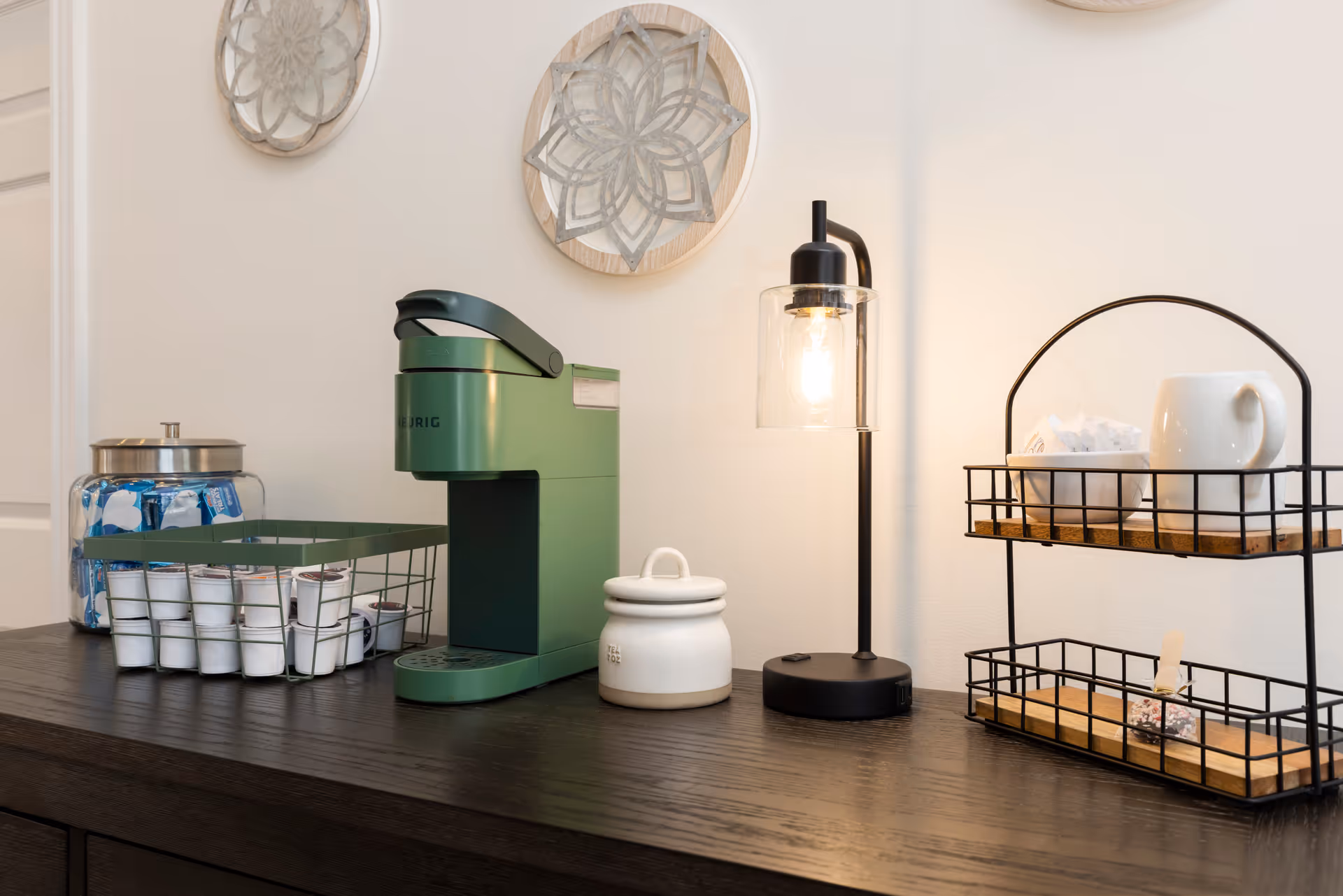 A countertop coffee station with a green single-serve coffee maker, coffee pods in wire baskets, a jar, a small lamp, and a two-tier metal rack against a light wall.