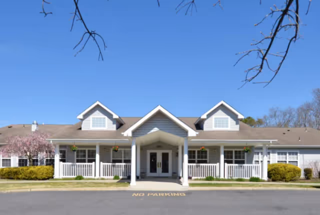 Front entrance of a single-story senior living building with a covered porch, white columns and dormer windows under a clear blue sky.